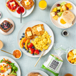 Load image into Gallery viewer, Assorted breakfast items including eggs, Dam Good English Muffins, fruit, and muffins on a light blue tablecloth.
