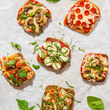 Load image into Gallery viewer, Assorted small Dam Good English Muffin pizzas on a white surface with fresh herbs.
